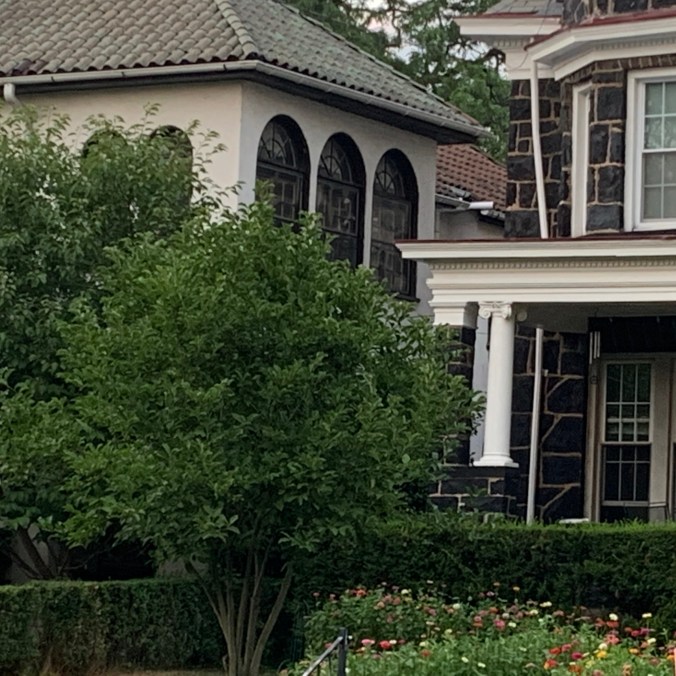 two old houses, one Spanish style and one made of large dark stone blocks, amid trees, hedges, and zinnias