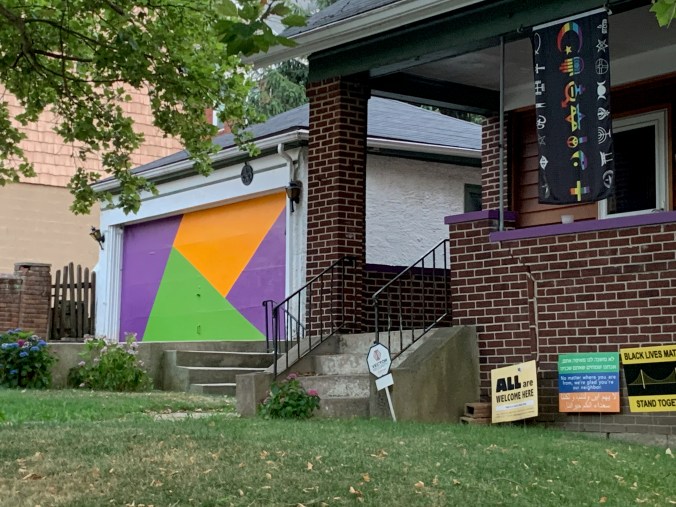 boring white garage with door painted in op-art blocks of purple, orange, and lime green; next to house with religious-inclusivity banner, several social-justice signs, and security-system sign