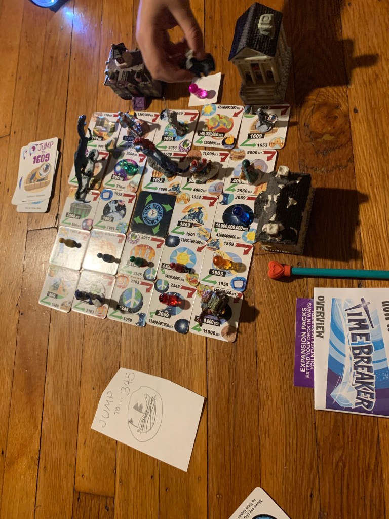 Full array of Time Breaker tiles, with the added tile and card nearby. Almost every tile is occupied by a playing piece--Time Breaker pawns, pawns from another game, small dinosaurs, and Christmas carolers. Three buildings from a miniature Christmas village are placed alongside. A child's hand is adding a figure to the scene.