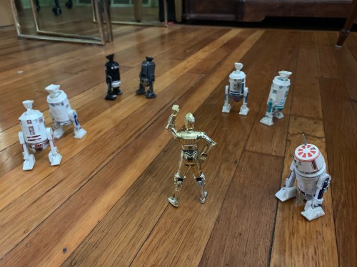 Star Wars action figures on a hardwood floor. C3-P0 has an authoritative stance with upraised arm. Six assorted R2 units are standing on their side legs with torso upside down and central foot in the air. R5-D4 is turned away toward the right.