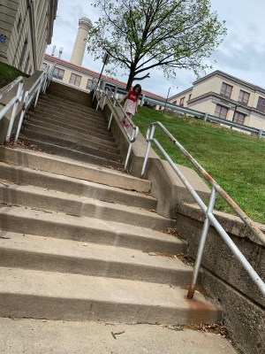 a little girl walking down the slanting concrete wall at the side of a tall staircase, holding onto the banister from the outside; a tree is just leafing out for spring
