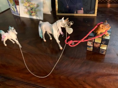 two toy unicorns connected by a necklace chain over their horns, with heart-shaped pendant dangling in front of the larger unicorn's face; a small bungee cord is hooked to the necklace and looped around the shoulders of a toy lion cub standing on top of a Rubik's cube; the whole scene is positioned in front of greeting cards and a portrait displayed on top of a wooden chest