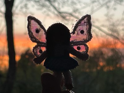 a tiny fairy doll held up to look out a window at the sunset; light is shining through her slightly frayed wings