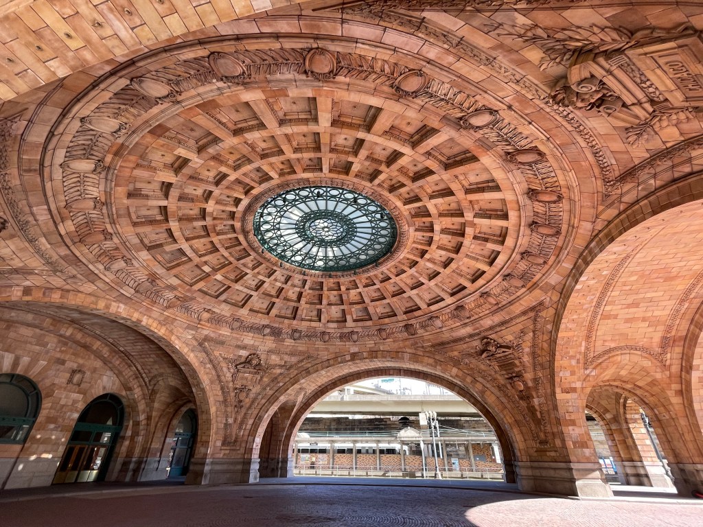 Round skylight with wrought-iron design in the center of a corbeled rotunda of reddish-brown blocks (are they brick or stone?) with leaf and shield motifs.  One of the arches around the perimeter opens into a 1980s-style light-rail station alongside a highway overpass.