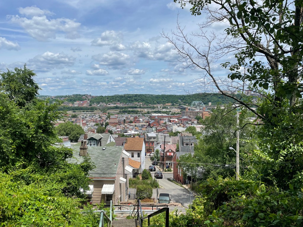 Looking between clumps of trees and down a concrete staircase, into a neighborhood of densely-spaced old houses; in the distance, churches, other buildings, an arched steel bridge, and across the river a hill with trees and buildings on it.