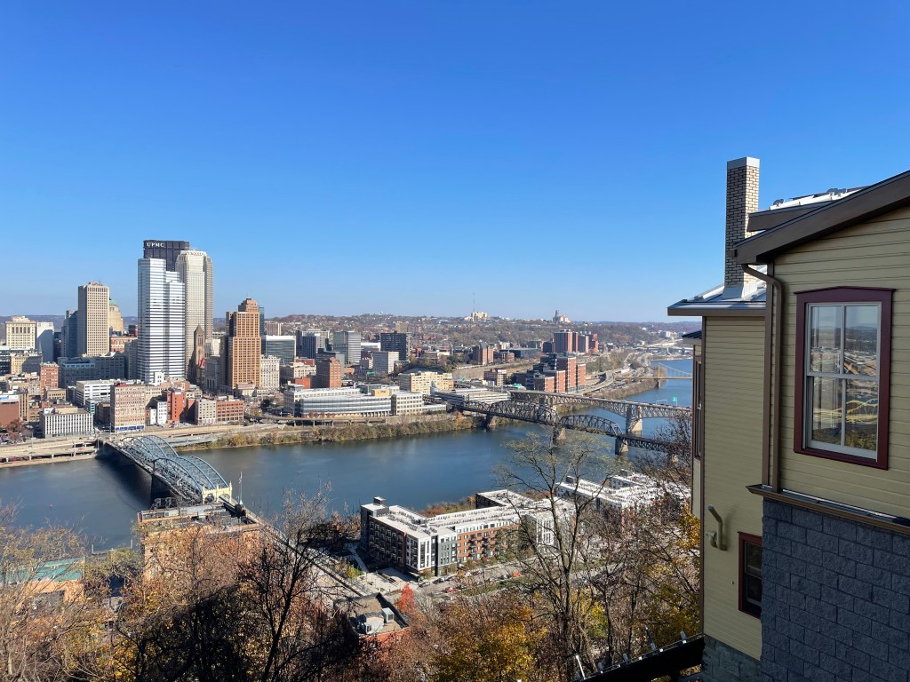 Bright blue sky above skyscrapers, river crossed by several bridges, big new apartment buildings on the near bank, all far below our vantage point.  A small building in the foreground at right is the upper station of Monogahela Incline, and a small amount of the incline track is visible descending the mountain.